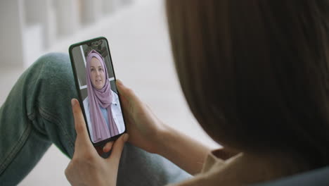 Frau-Nutzt-Medizinische-App-Auf-Dem-Smartphone-Und-Berät-Sich-Per-Videokonferenz-Mit-Einer-Arabischen-Muslimischen-Frau-Im-Hijab-Arzt.-Frau-Nutzt-Online-Chat,-Um-Mit-Familientherapeuten-Und-Einer-Coronavirus-Pandemie-Zu-Sprechen.